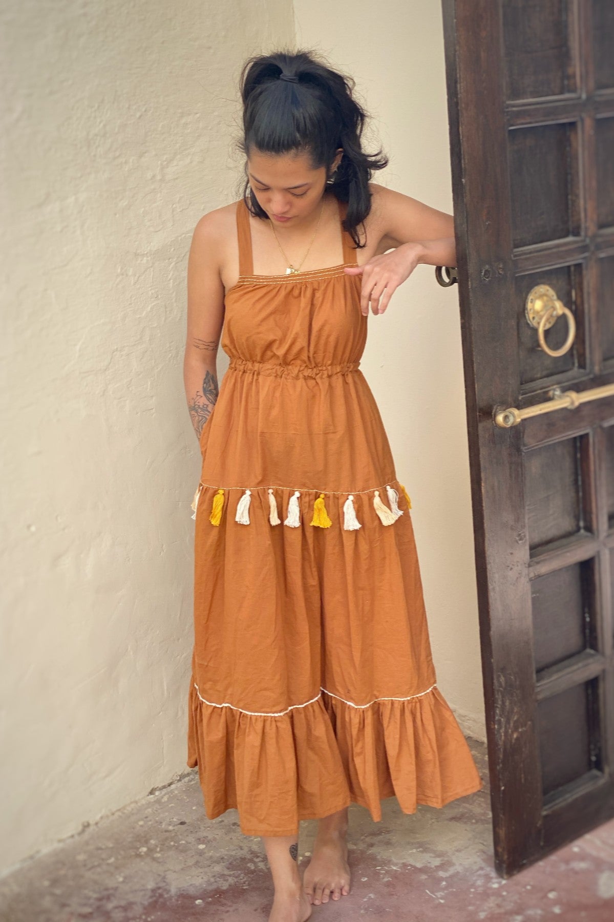 Woman dressed in a clay coloured, full length tiered cotton dress with yellow, orange and white hand embroidered details and tassels. 